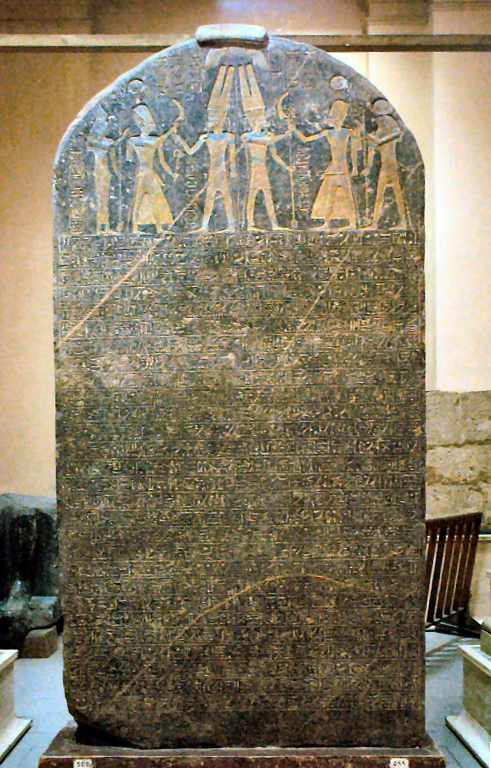 The Merneptah Stele in the Egyptian Museum, Cairo — a tall round-topped black granite slab covered in hieroglyphic text, with carved scenes of gods and the king across the top.

