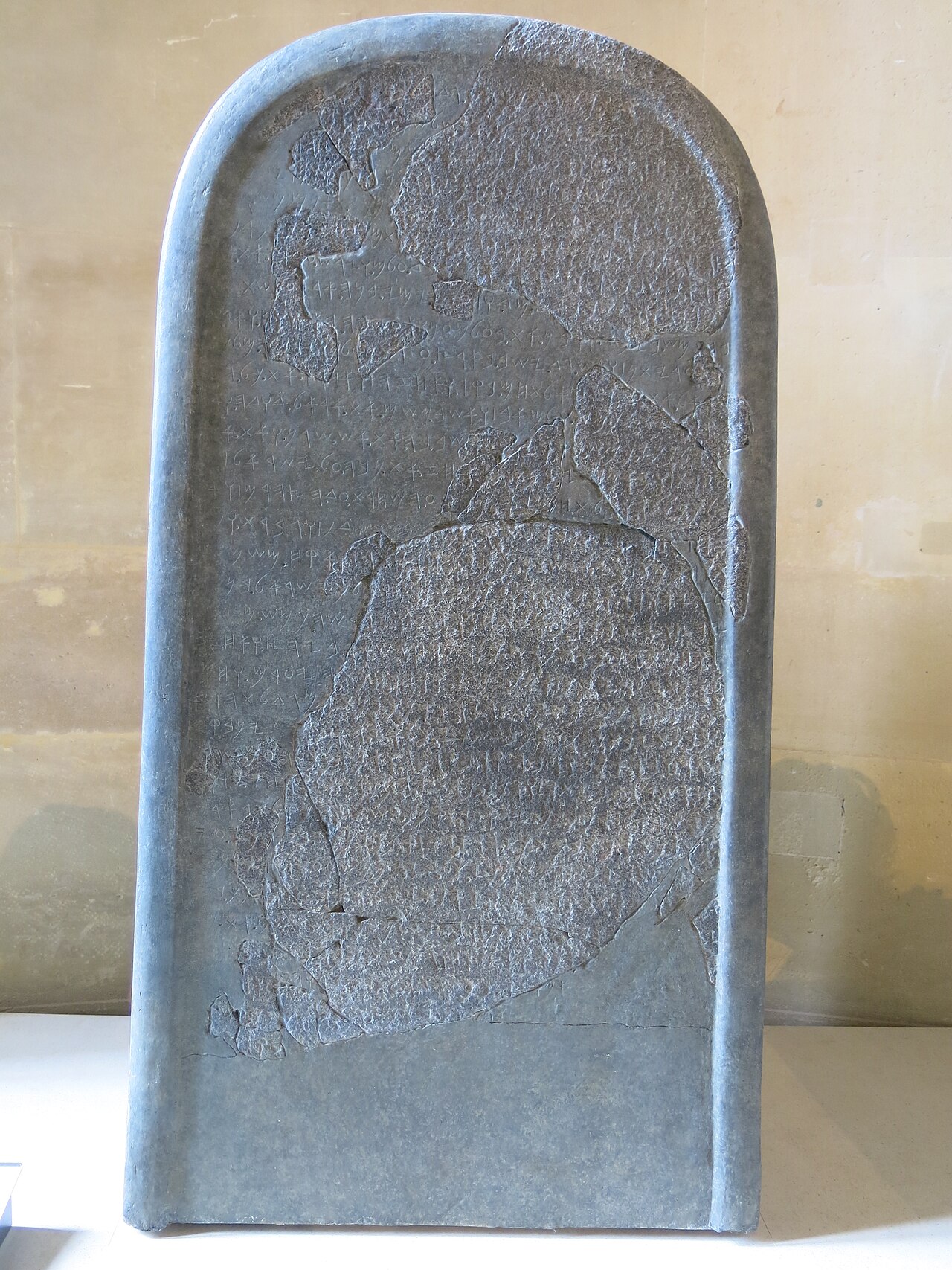 The Mesha Stele in the Louvre — a rounded-top black basalt slab reassembled from fragments, its face covered in lines of early alphabetic Moabite inscription.