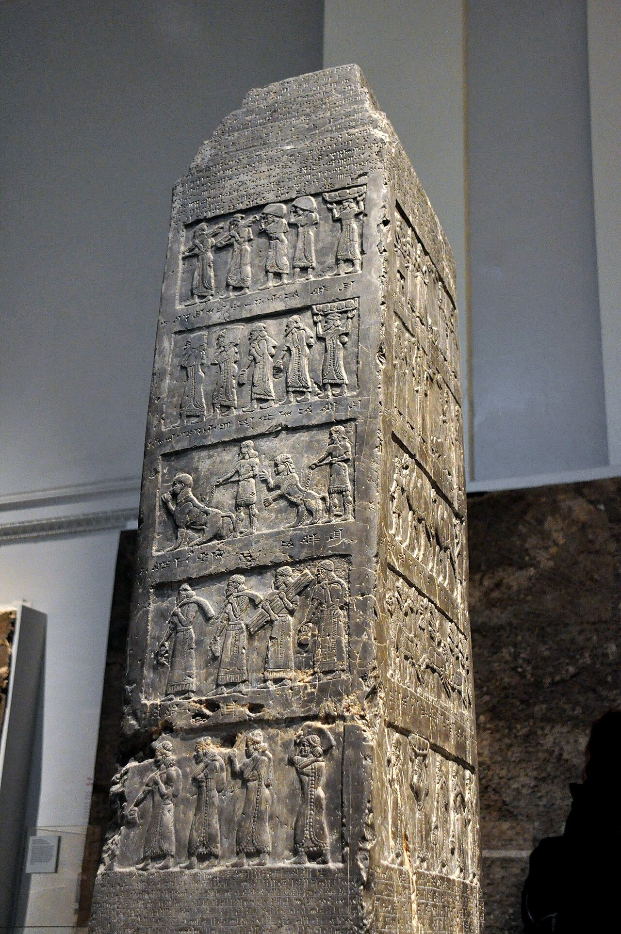 The Black Obelisk of Shalmaneser III in the British Museum — a four-sided black limestone pillar crowned like a ziggurat, its faces carved with registers of tribute-bearing figures and cuneiform captions.