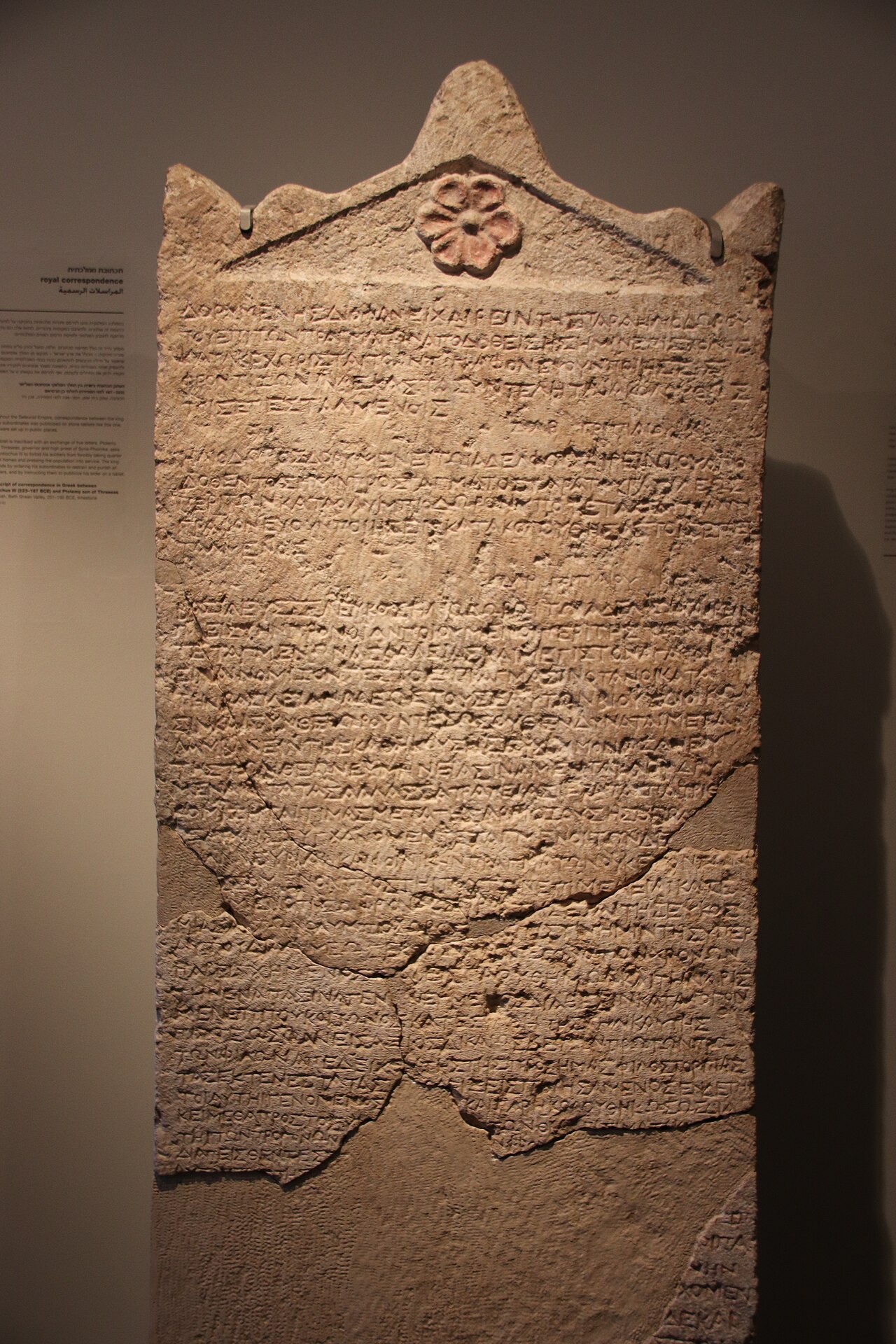 The Heliodorus Stele at the Israel Museum — a tall limestone slab with a rounded top, its face carved with many lines of even Greek letters, the lower part restored from excavated fragments.
