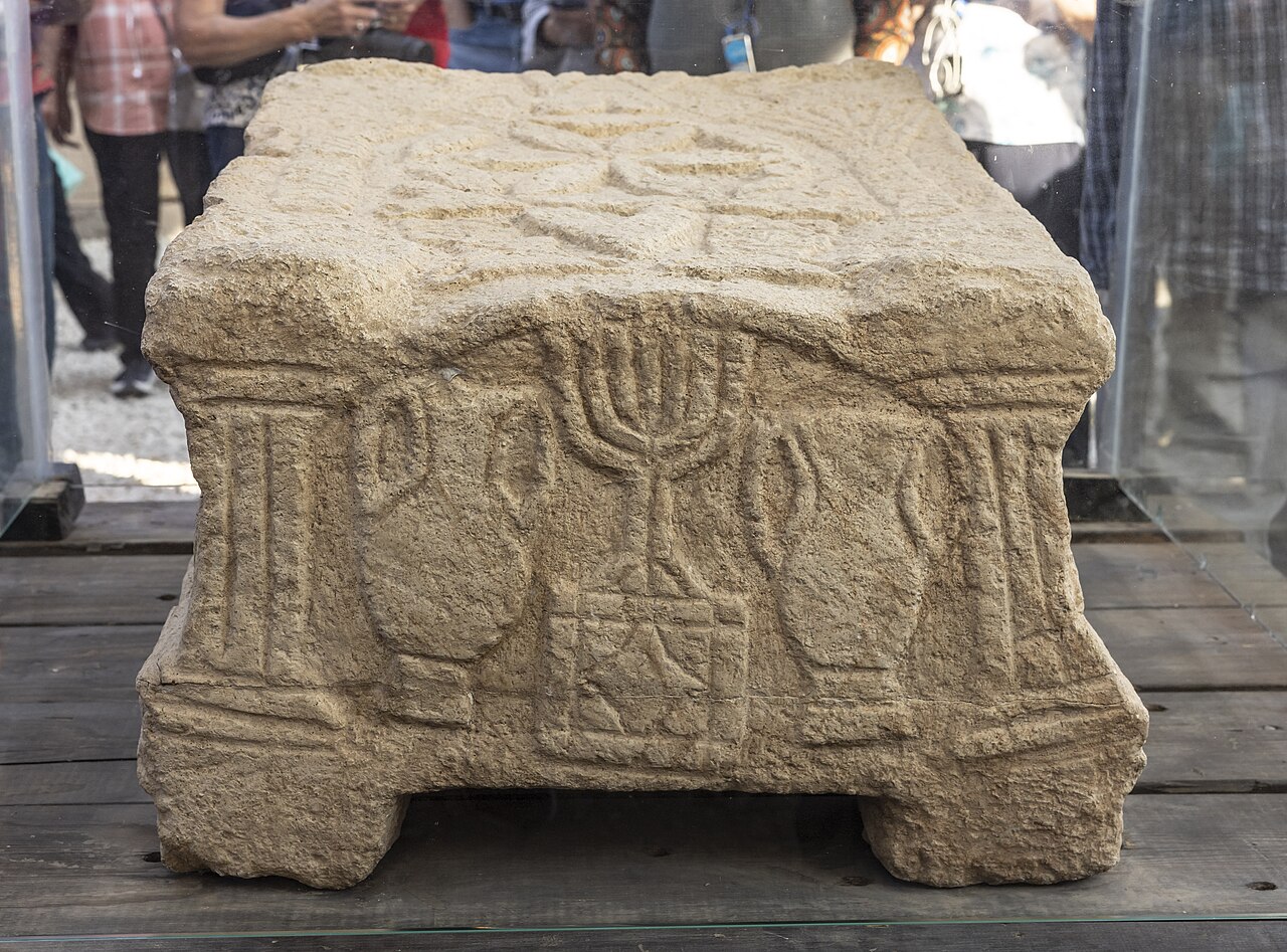 The Magdala Stone displayed in a glass case at the Magdala site — a carved limestone block on four squat feet, its front face bearing a seven-branched menorah in relief between columns and amphorae.
