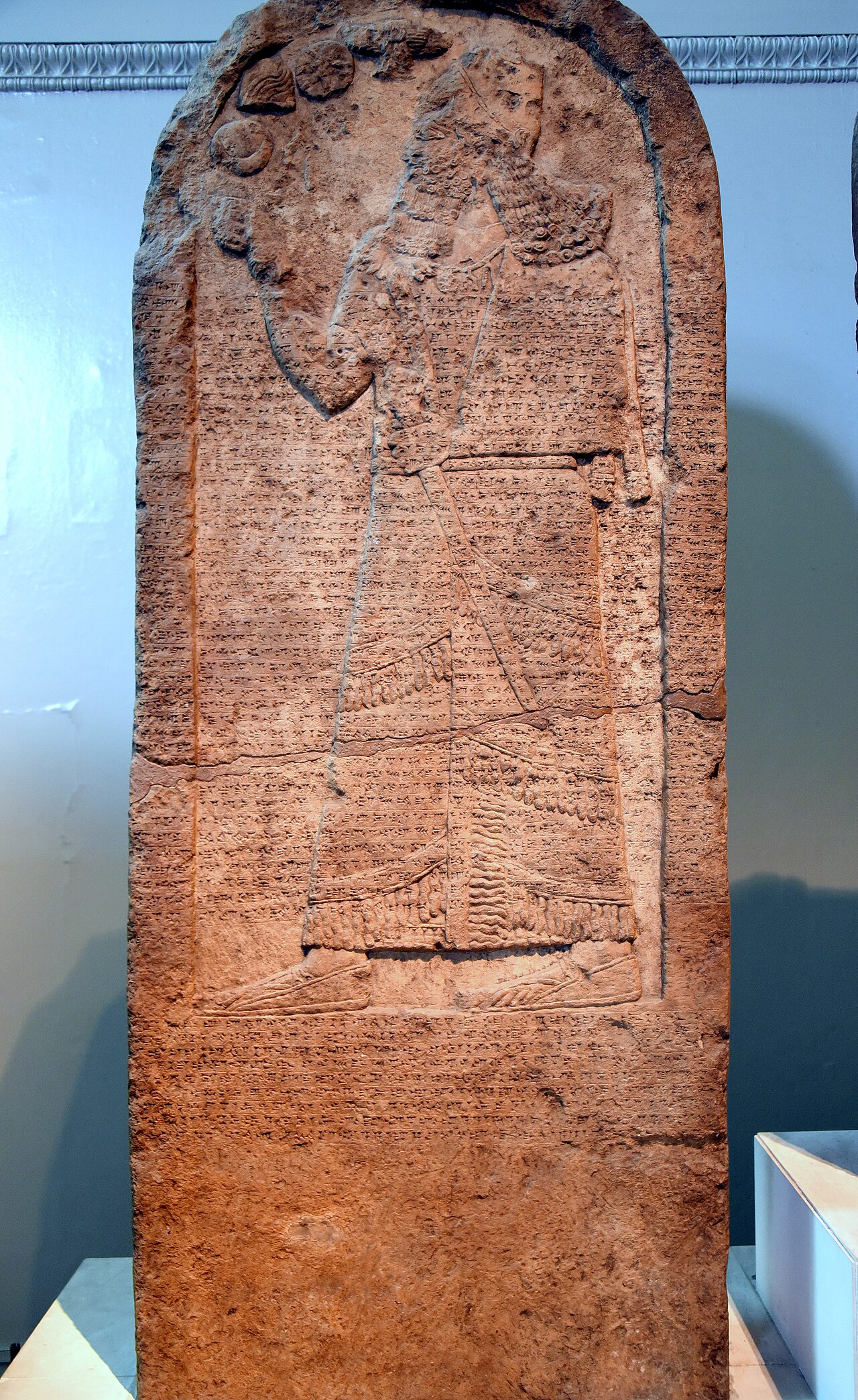 The Kurkh Monolith in the British Museum — a rounded-top limestone stele carved with a standing figure of Shalmaneser III before divine symbols, the surface covered in cuneiform annals.
