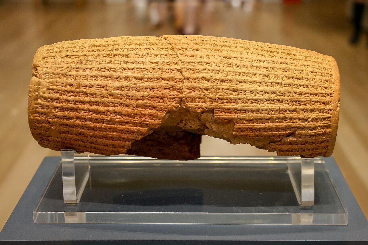 The Cyrus Cylinder in the British Museum — a barrel-shaped clay cylinder covered in lines of cuneiform, displayed in a glass case.
