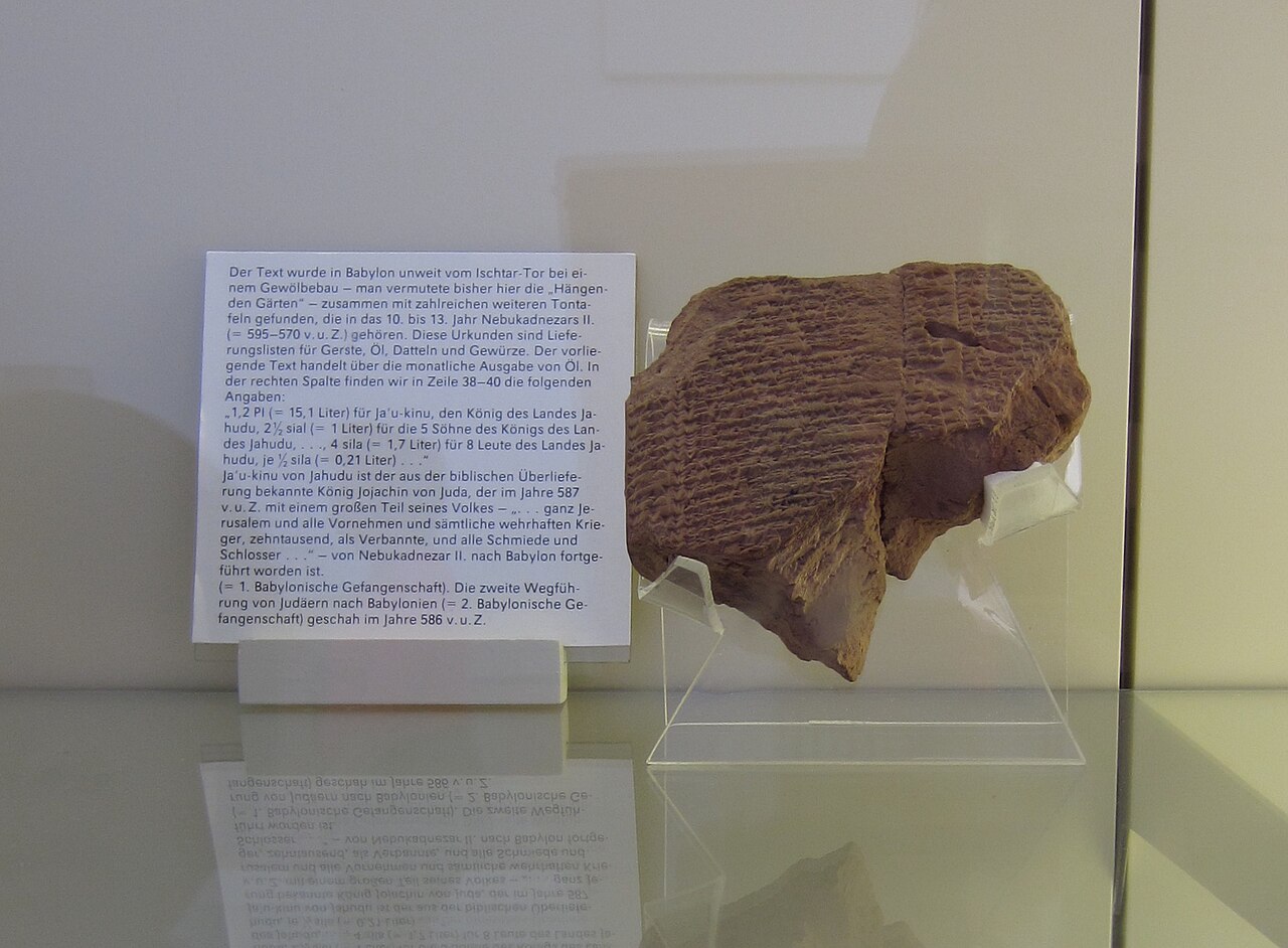One of the Jehoiachin ration tablets in the Pergamon Museum, Berlin — a fragmentary clay tablet of cuneiform accounting entries, displayed beside the museum's label card.

