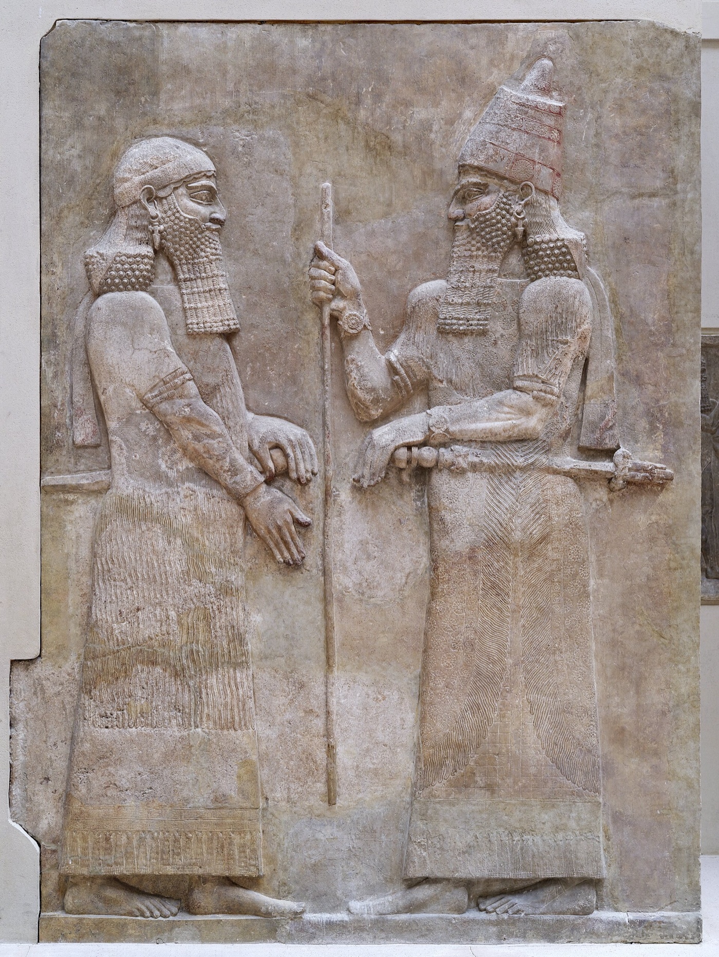 Wall relief from Sargon II's palace at Khorsabad, in the Louvre — the king, in a tall tiara and holding a long staff, faces a beardless high dignitary; both figures stand in profile in finely carved alabaster.
