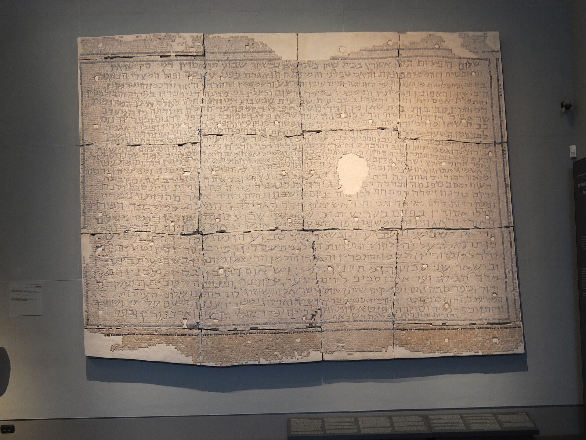 The Rehob halakhic mosaic on display at the Israel Museum — a large white mosaic panel filled with lines of black Hebrew lettering.
