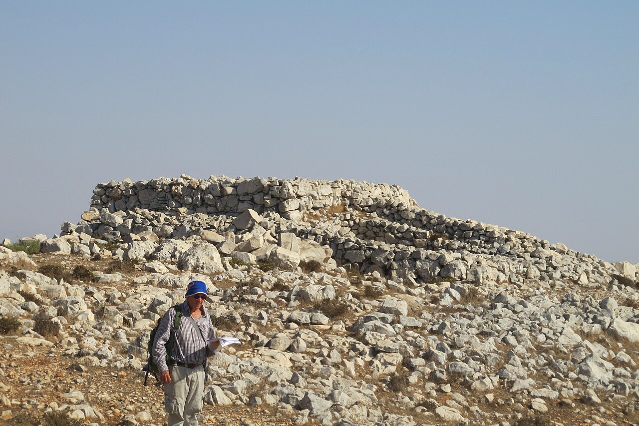 The main structure of the Mount Ebal site — a broad rectangular platform of unhewn field stones rising from a stony slope on Har Eival, with a hiker standing on the rocky ground before it.