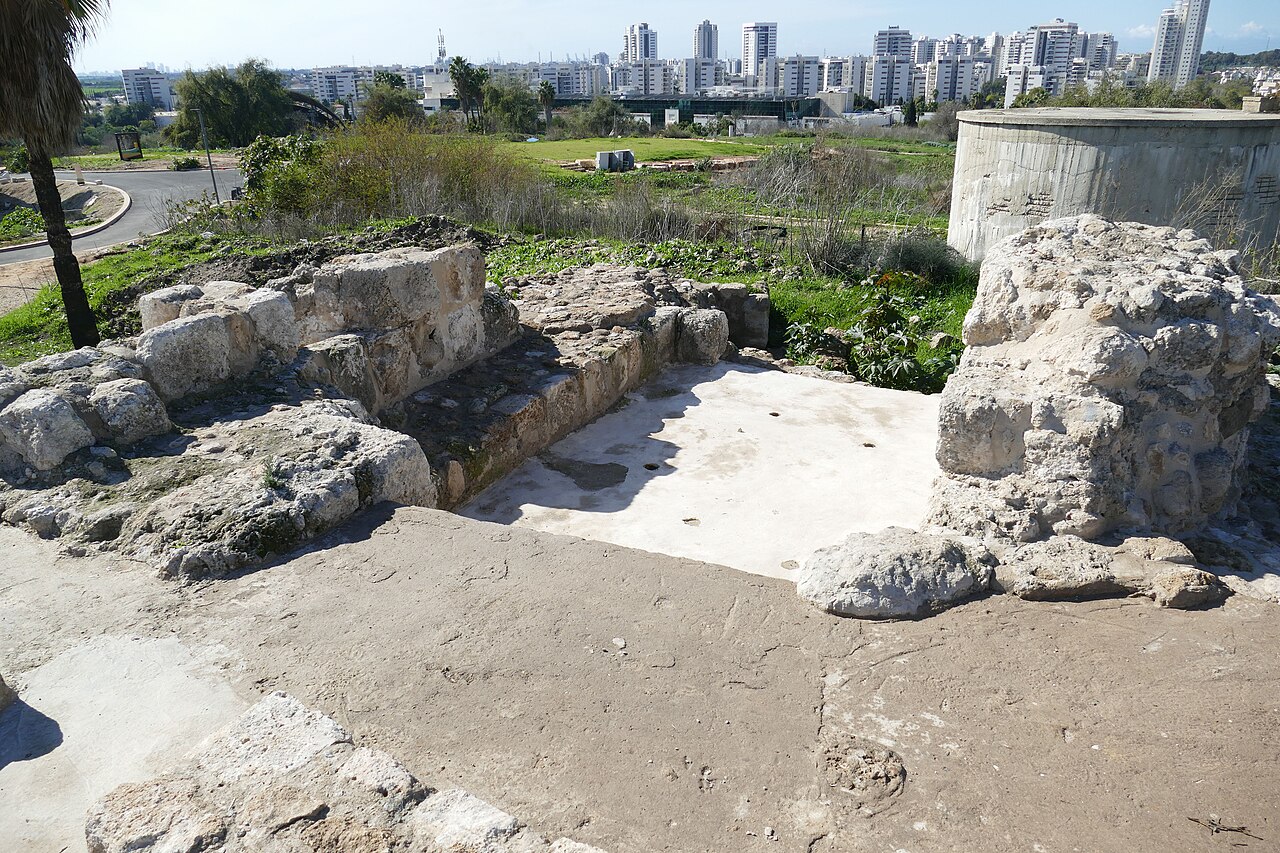 Excavated stone ruins on Tel Yavne, the mound of ancient Yavneh, with the apartment towers of the modern city of Yavne rising on the coastal plain behind.
