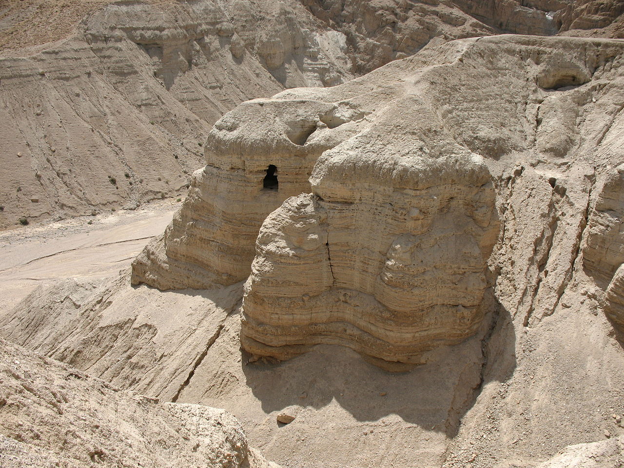 Cave 4 at Qumran — the collapsed marl cliff spur with its dark cave openings above the dry wadi, the single richest of the eleven scroll caves, which yielded thousands of Dead Sea Scroll fragments in 1952.
