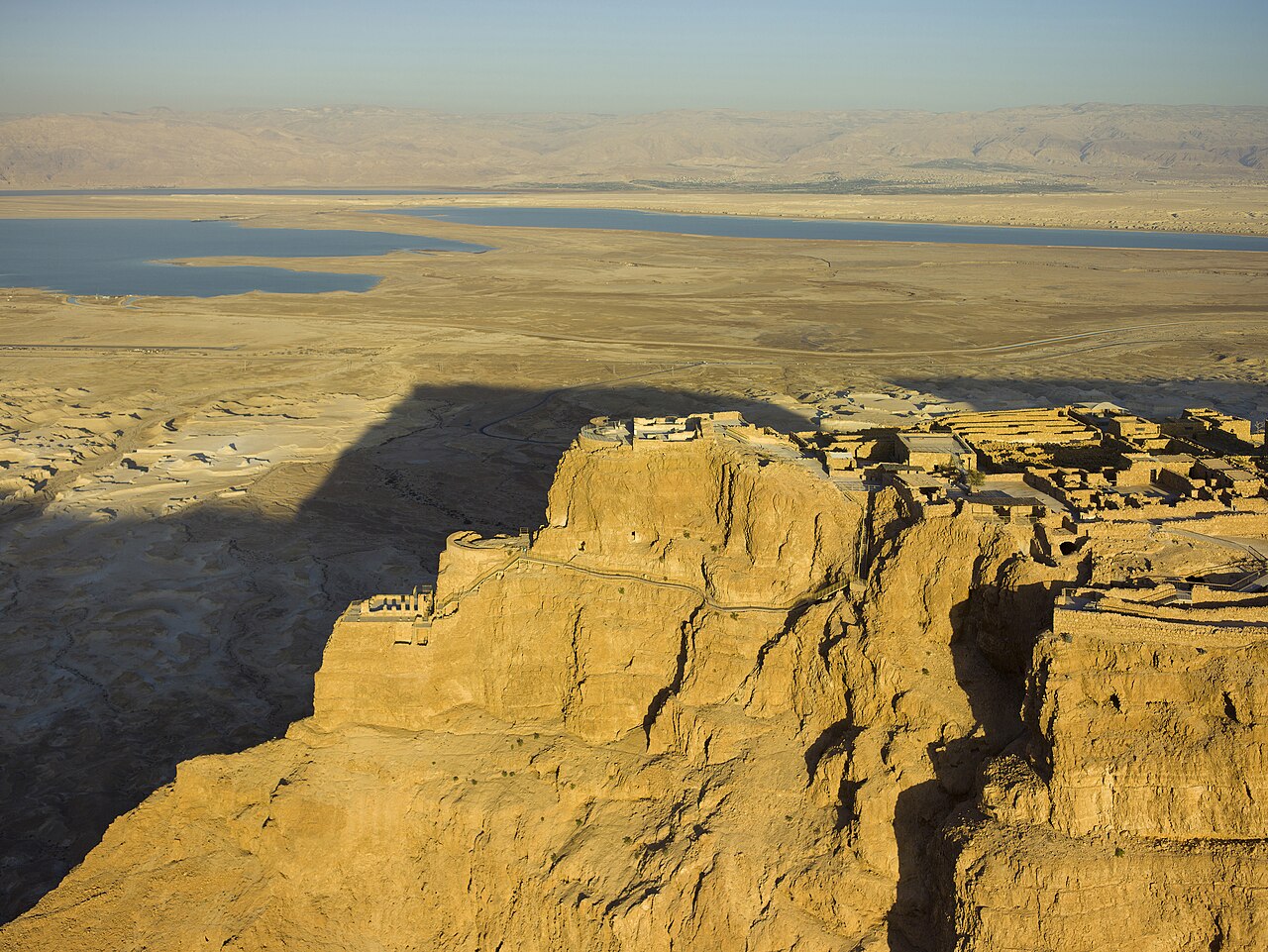 Aerial view of Masada at sunset — the isolated flat-topped mesa with Herod's Northern Palace terraces at its tip and the ruins on the summit, the Dead Sea and the mountains of Moav in the distance.
