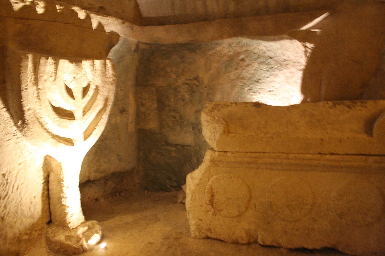 Inside Catacomb no. 20 at Beit Shearim — a great seven-branched menorah carved in relief from the living rock beside a stone sarcophagus (part of the menorah's central shaft is a modern restoration).