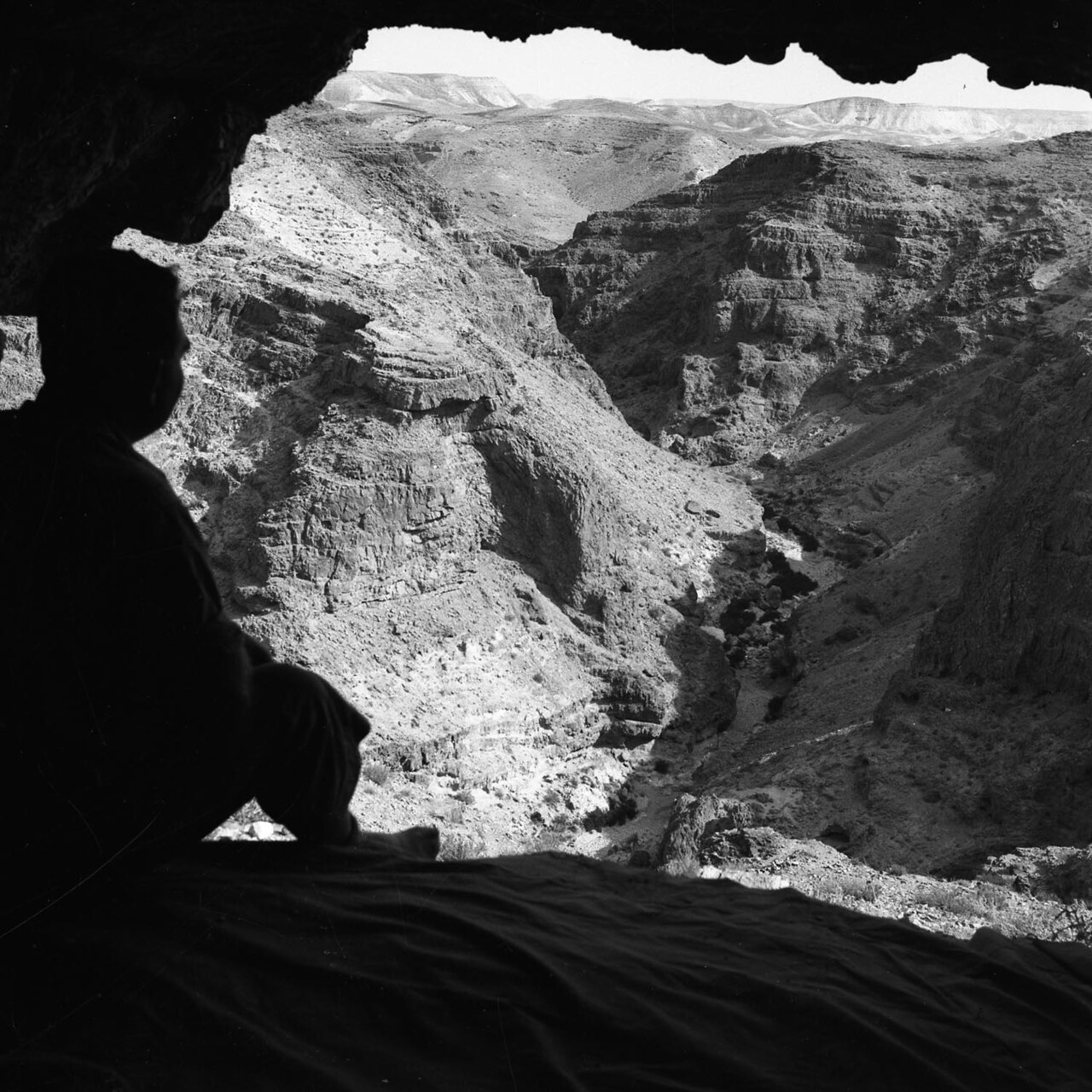 Looking out of the mouth of the Cave of Letters — a figure from the 1950s expeditions silhouetted against the sheer cliffs and dry gorge of the Nahal Hever canyon below.

