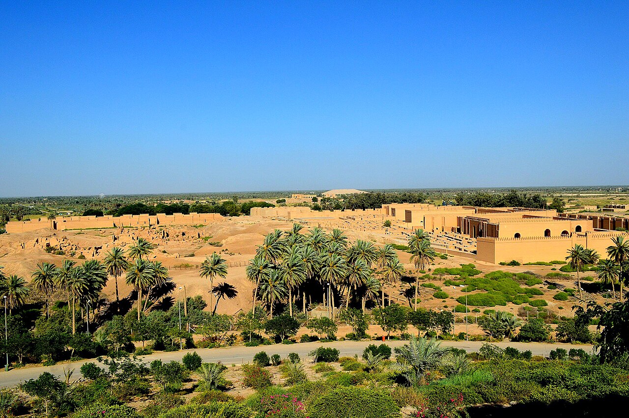 The ruins of ancient Babylon near Hillah, Iraq — excavated mounds and mud-brick remains among date palms, with the modern reconstructions of the city's palace walls (built over the ruins in the twentieth century) at right.
