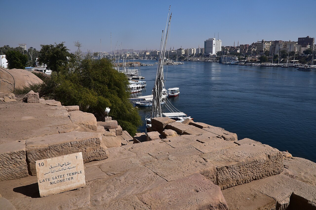 The southern end of Elephantine Island at Aswan — ancient stone ruins by the late Satet temple nilometer in the foreground, feluccas moored on the Nile, and the Aswan corniche across the water. The Judean colony's quarter stood among these ruins.