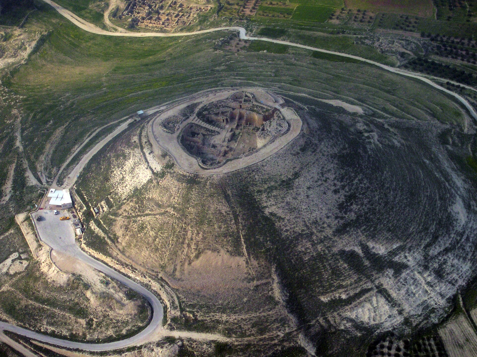Aerial view of Herodium, an artificial cone-shaped hill with the excavated circular palace-fortress sunk into its flattened summit, surrounded by fields and desert roads.
