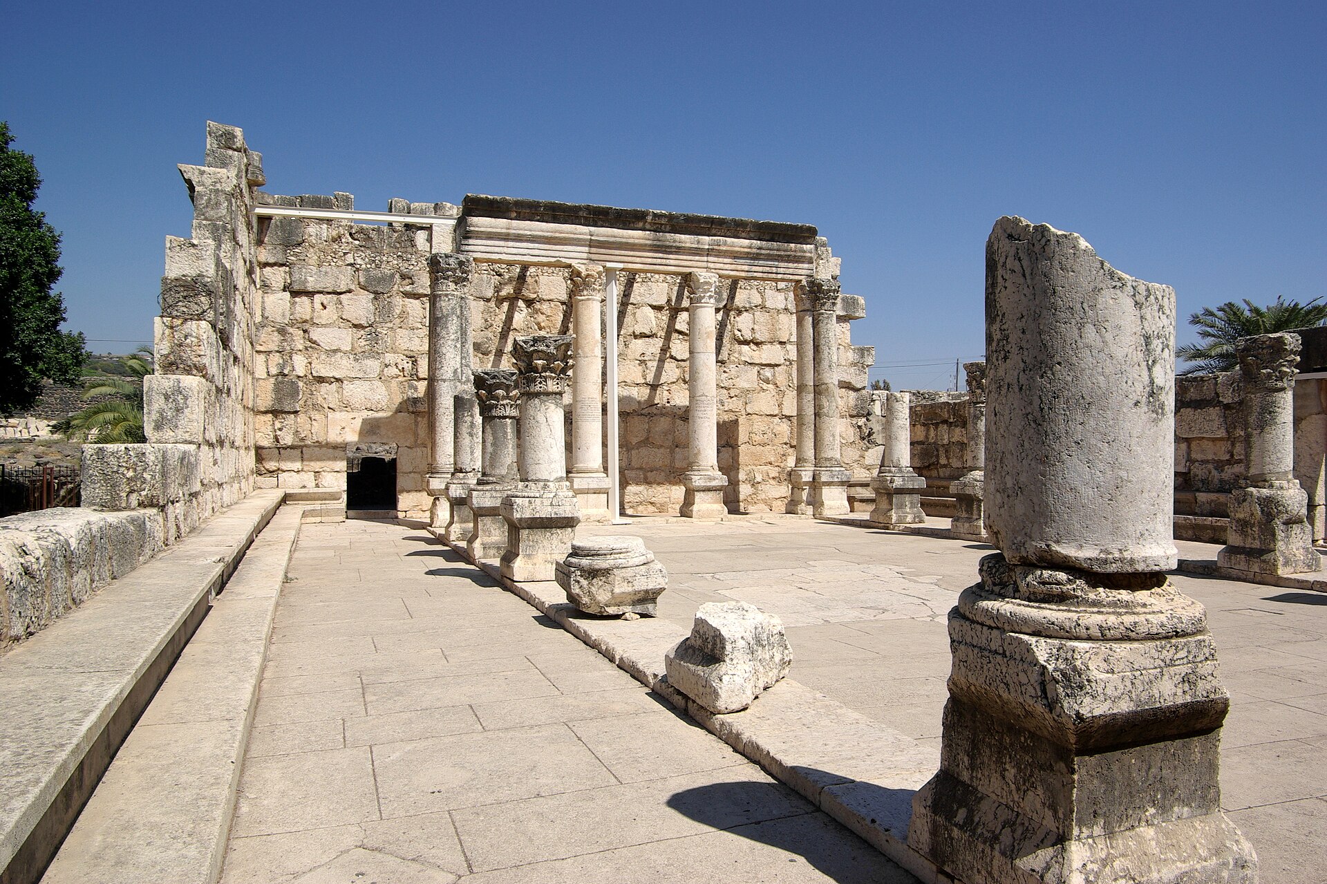The white limestone synagogue of Capernaum — a ruined colonnaded prayer hall with Corinthian columns, carved lintels, and stone benches — under a deep blue Galilee sky.
