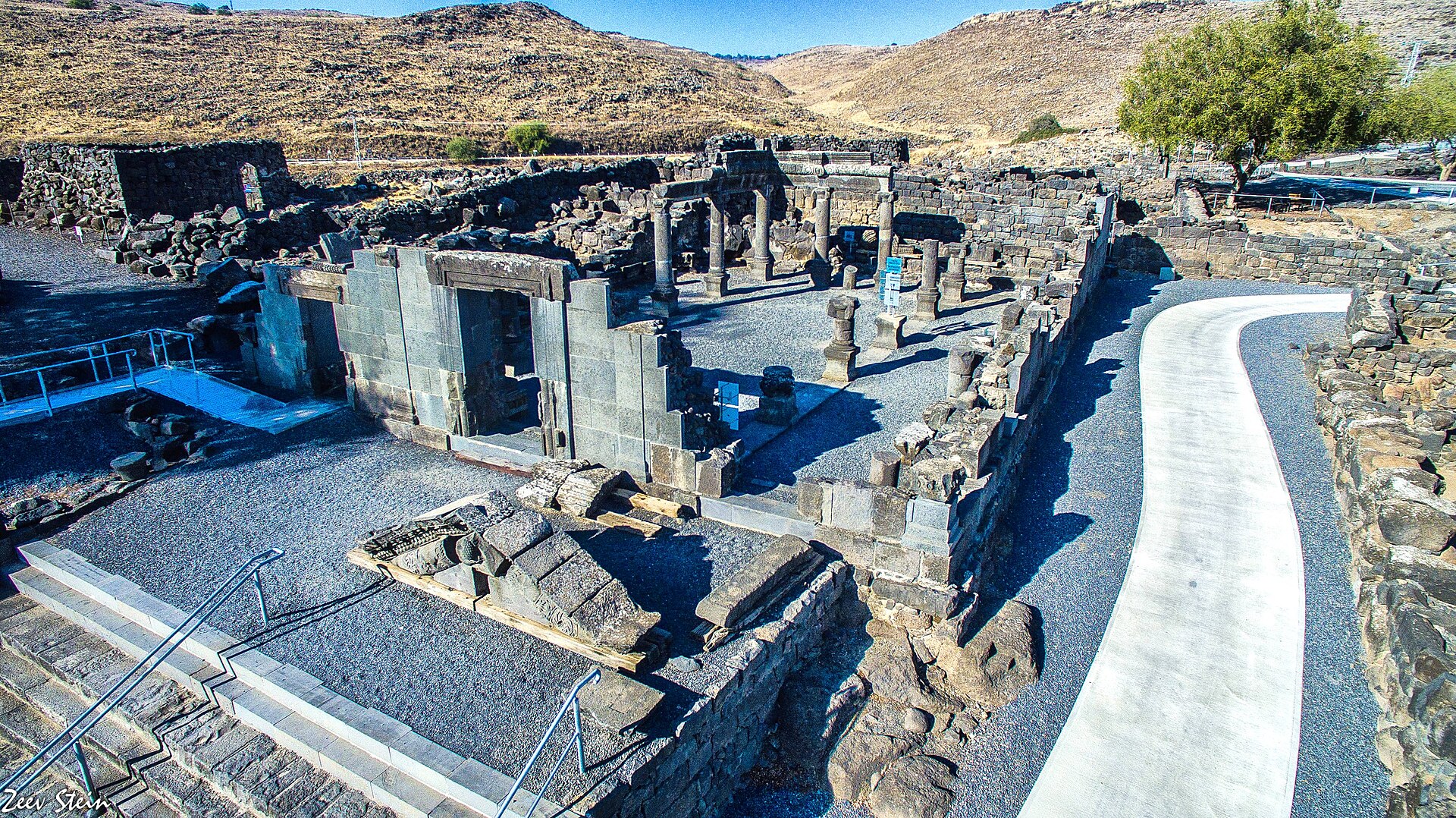 The black basalt synagogue of Korazim seen from above — carved doorframes, column stumps, and re-erected columns amid the ruins of the basalt town, with the dry hills of the plateau behind.

