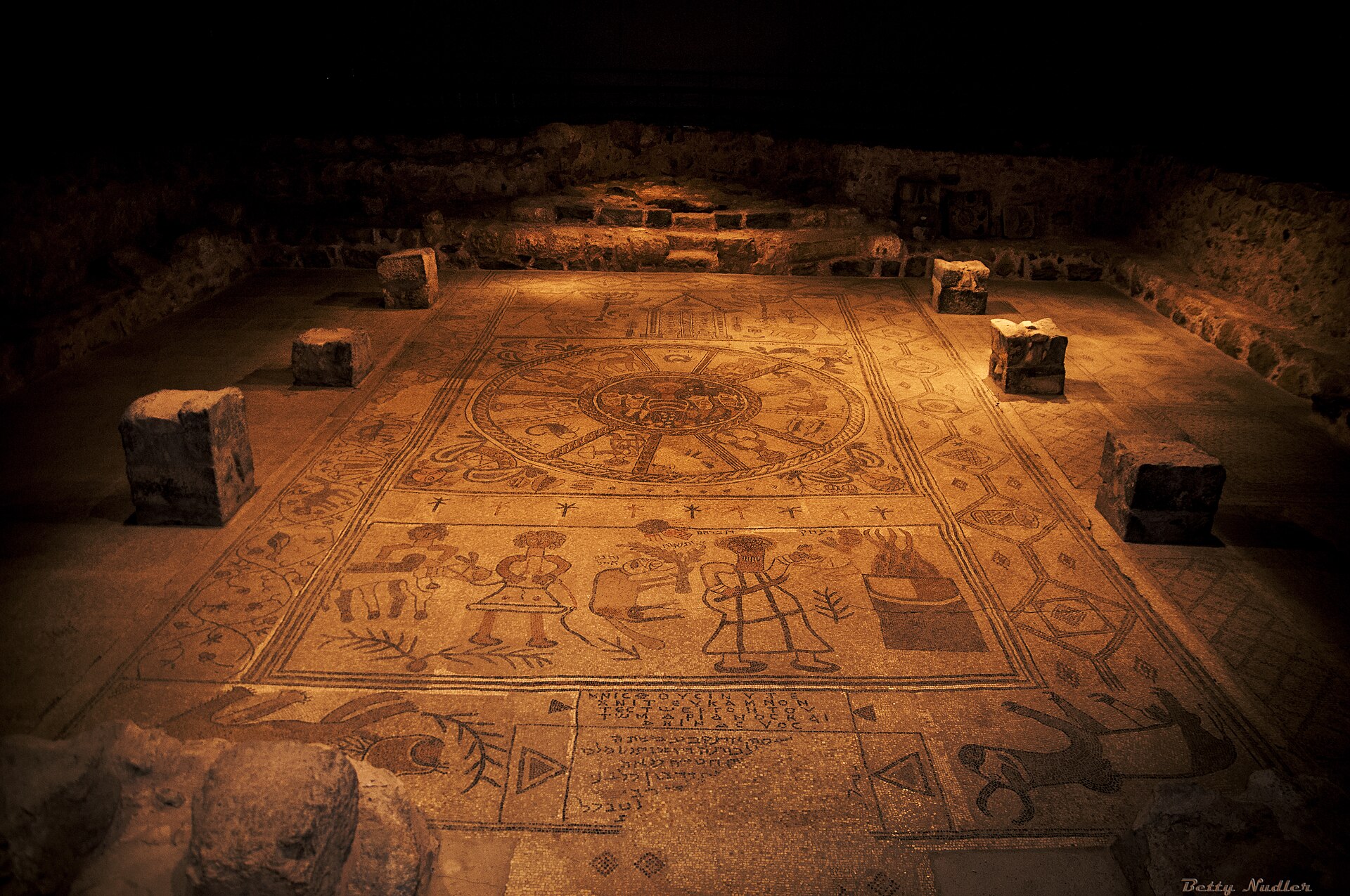 The Beit Alpha synagogue mosaic floor seen under evening lighting inside its shelter — the zodiac wheel at center, the binding of Yitzchak panel in the foreground, and column bases around the nave.
