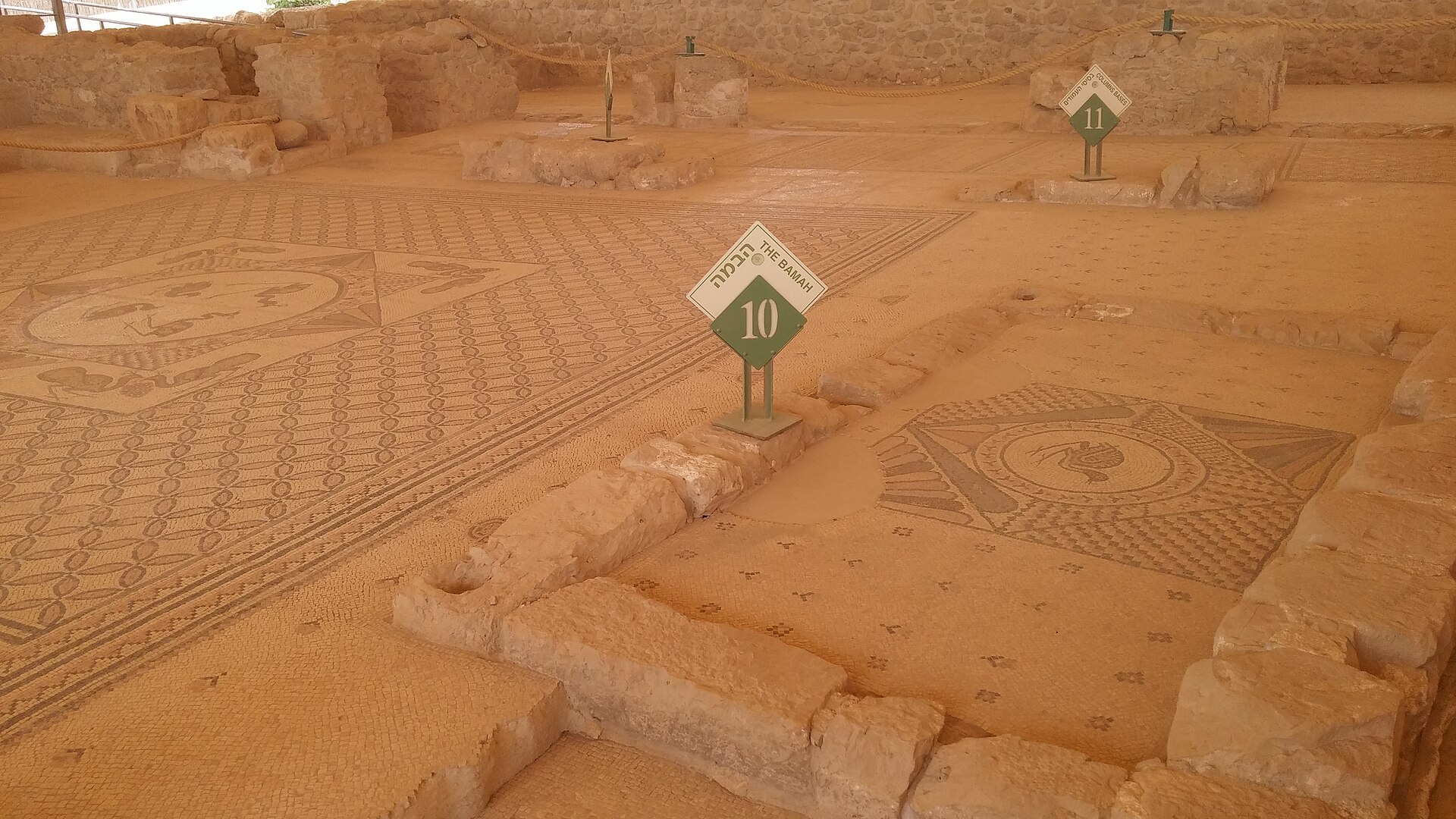 The mosaic floors of the ancient Ein Gedi synagogue under a protective shelter, with geometric carpets, a bird medallion, and a numbered sign marking the bamah.
