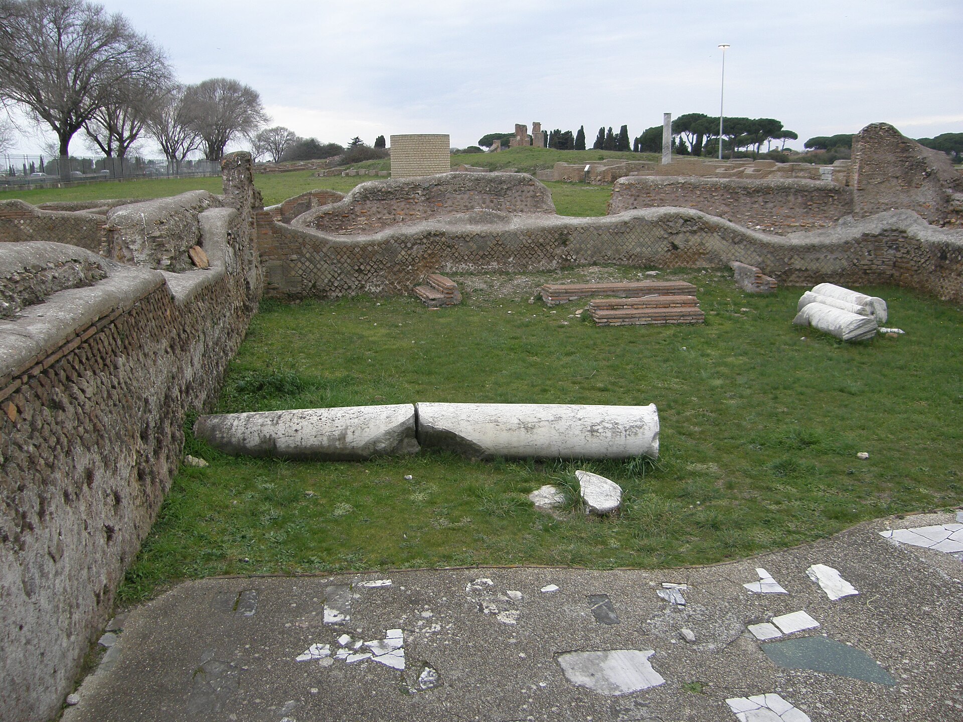 The ruins of the Ostia synagogue's interior — brick walls and re-erected grey marble columns of the assembly hall, open to the sky among the pines of Ostia Antica.
