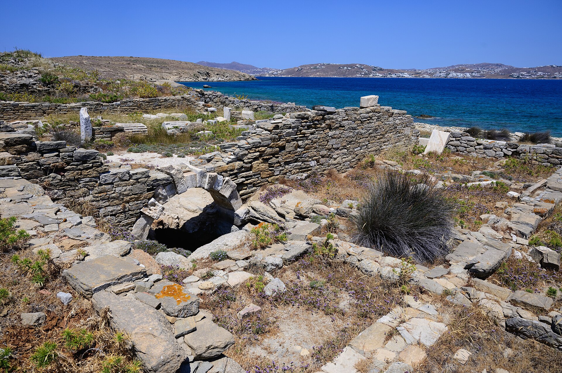 The low ruined walls and scattered marble of building GD 80, the candidate synagogue on the eastern shore of Delos, with the sea behind.
