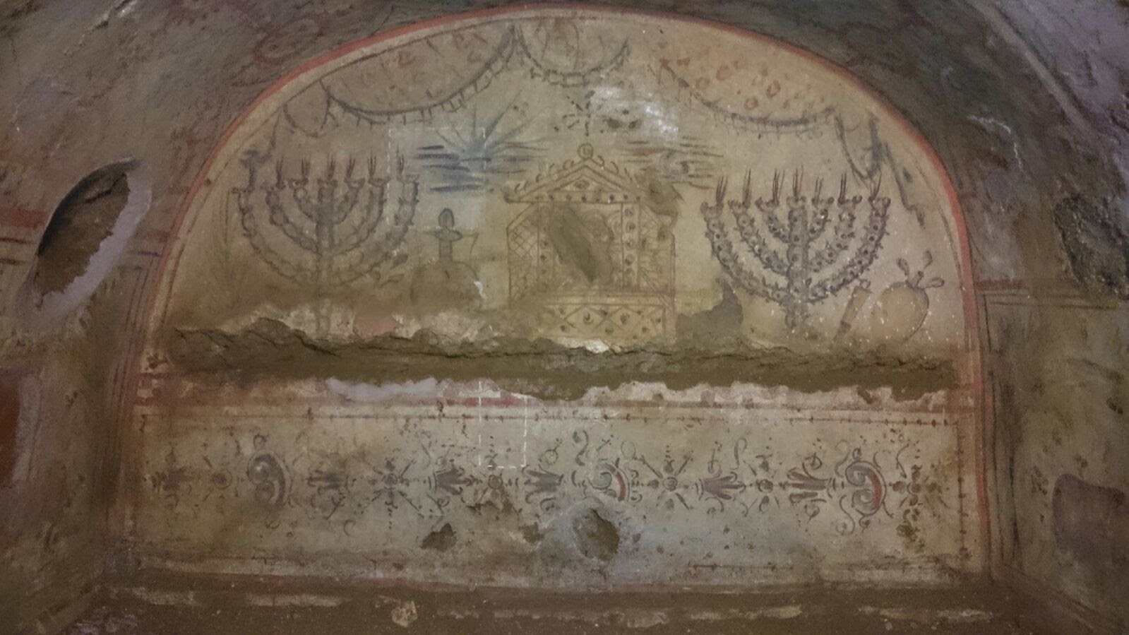 The painted arcosolium of the Villa Torlonia catacombs — an open Torah shrine with scrolls visible inside, flanked by two large menorot, with etrog, shofar, and oil flask, over a band of floral ornament.

